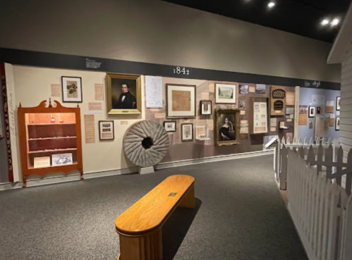 A museum exhibit featuring framed artworks, historical documents, and a wooden bench in a well-lit space.