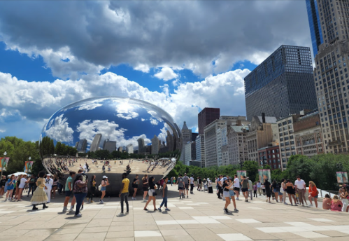 A large reflective sculpture in a park, surrounded by people and city buildings under a partly cloudy sky.