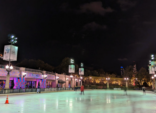 A nighttime ice skating rink with colorful lights, surrounded by trees and buildings in the background.