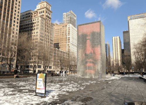 A large digital installation displays a face, surrounded by a snowy urban park with skyscrapers in the background.