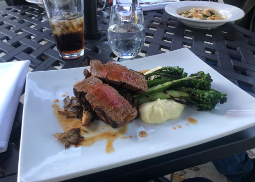 A plate of sliced beef with a red center, served with mashed potatoes and broccolini, alongside drinks on a table.