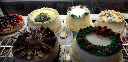 A display of various decorated cakes, including fruit-topped, festive, and chocolate varieties.