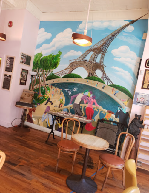A colorful mural featuring the Eiffel Tower and people enjoying a park scene, with wooden chairs and a table in the foreground.