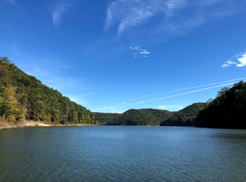 A serene lake surrounded by green hills under a clear blue sky with wispy clouds.