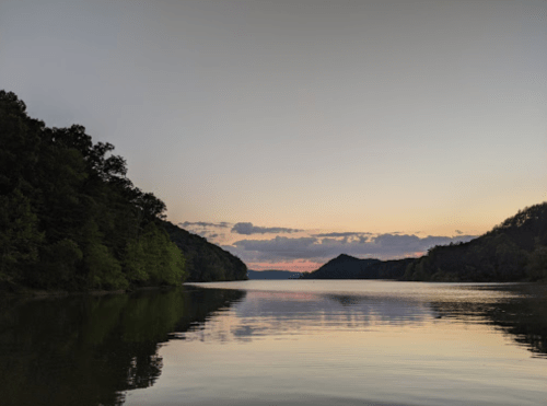 A serene river scene at sunset, with calm waters reflecting the colorful sky and lush green hills on either side.