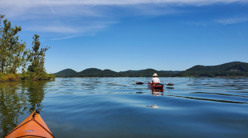 A person in a kayak paddles on a calm lake, surrounded by trees and distant hills under a clear blue sky.