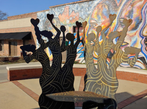 A metal sculpture of figures with raised arms and hearts, set against a colorful mural backdrop.