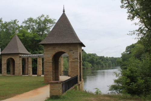 A stone gazebo with pointed roofs overlooks a calm river, surrounded by lush greenery and a cloudy sky.
