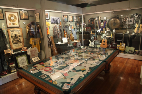 A music museum display featuring guitars, memorabilia, and framed records in a glass-topped showcase.