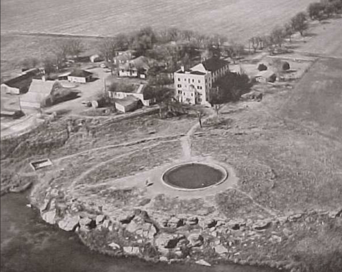 Aerial view of a rural area featuring a large building, small structures, and a circular pond surrounded by land.