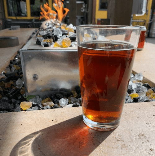A glass of amber-colored drink sits on a table beside a fire feature with colorful stones.