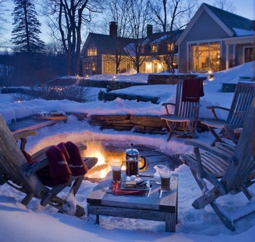 Cozy outdoor scene with a fire pit surrounded by wooden chairs, snow, and a warm house in the background at dusk.