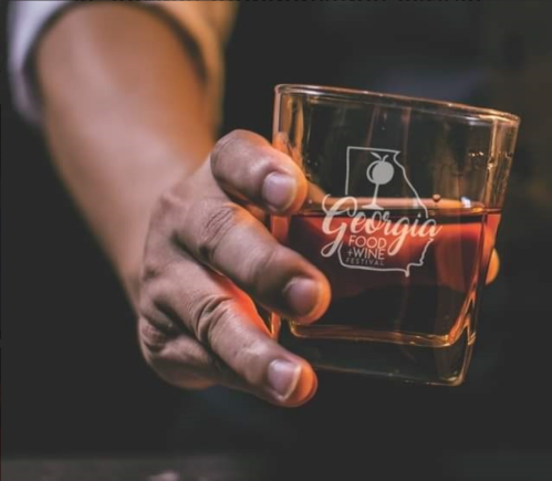 A hand holding a glass with the Georgia Food & Wine Festival logo, filled with a dark beverage.