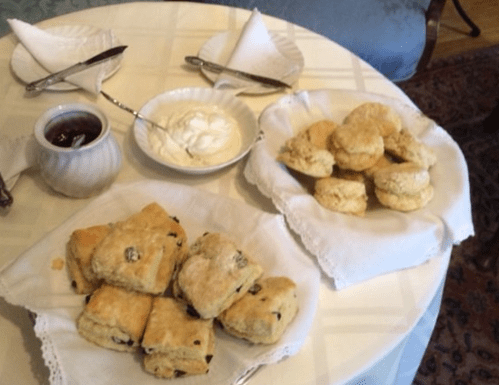 A table set with two plates of scones, one with currants, accompanied by cream and jam in small bowls.