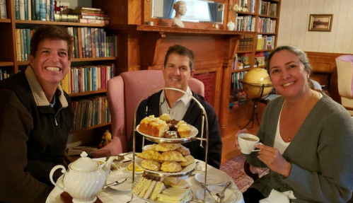 Three people enjoy tea and pastries at a cozy table, surrounded by books and a warm, inviting atmosphere.