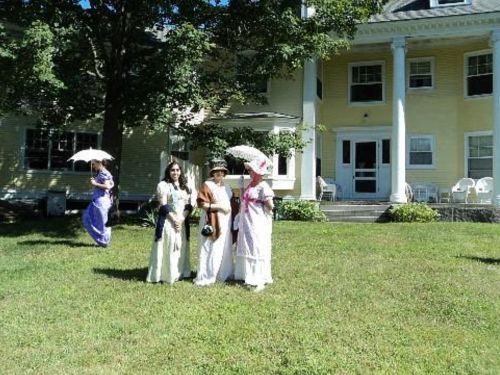 A group of women in historical dresses holding parasols, standing on a lawn in front of a yellow house.