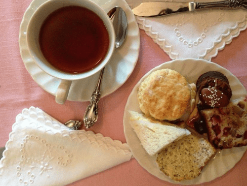 A cup of tea beside a plate of assorted pastries, including scones, cookies, and slices of cake, on a pink tablecloth.