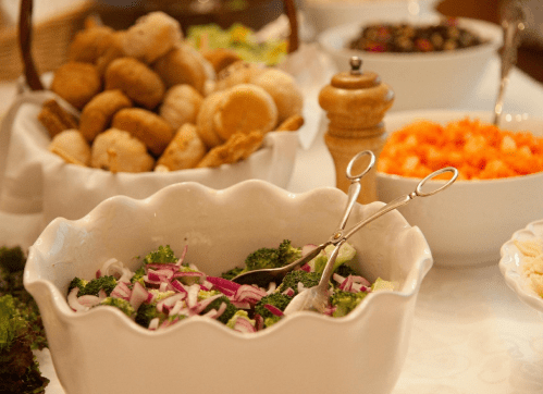 A table spread with various dishes, including a broccoli salad, rolls, and a bowl of carrots.