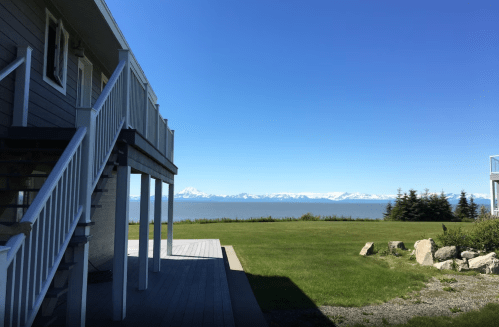 A view from a house with a deck overlooking a grassy area and a calm sea, with mountains in the distance under a clear blue sky.