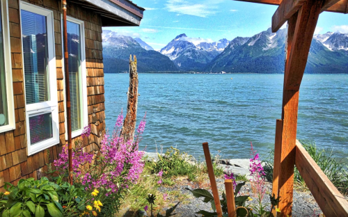 Scenic view of mountains and water from a wooden porch, with colorful flowers in the foreground.