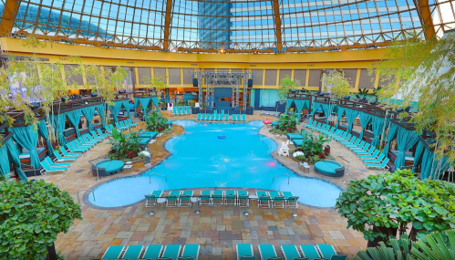 Indoor pool area with lush greenery, lounge chairs, and a large skylight overhead. Bright and inviting atmosphere.