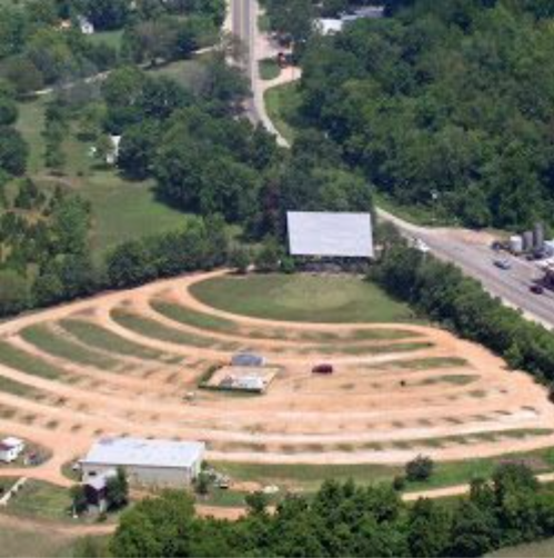 Aerial view of a rural area with winding dirt paths, green trees, and a few buildings near a road.