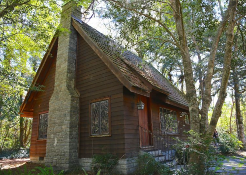 A quaint wooden cabin surrounded by trees, featuring a stone chimney and decorative windows.