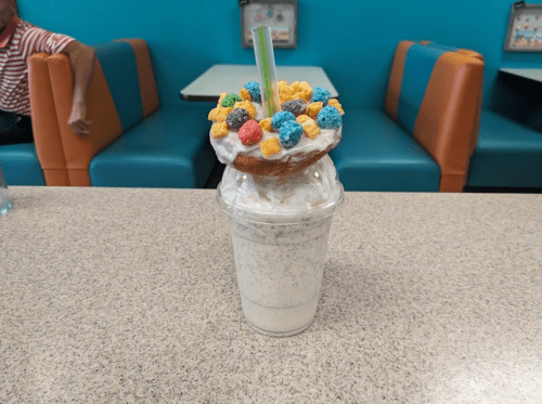 A milkshake topped with whipped cream, a donut, and colorful cereal, sitting on a table in a diner.