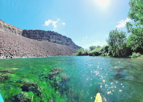 A serene river scene with clear green water, rocky cliffs, and lush trees under a bright blue sky.