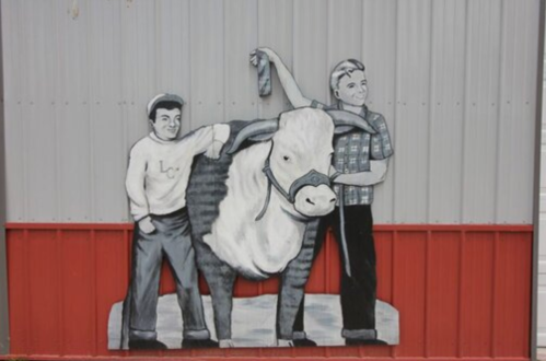 Two men stand beside a large bull, one holding a ribbon, in a black-and-white mural on a barn wall.