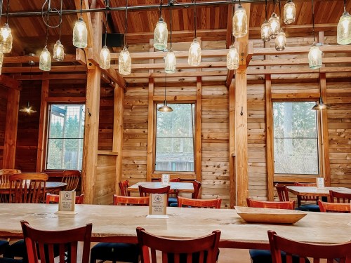 Cozy wooden restaurant interior with tables, chairs, and hanging mason jar lights. Large windows let in natural light.