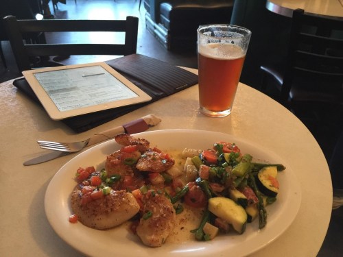 A plate of sautéed scallops with vegetables, an iPad, and a glass of amber beer on a table in a casual setting.