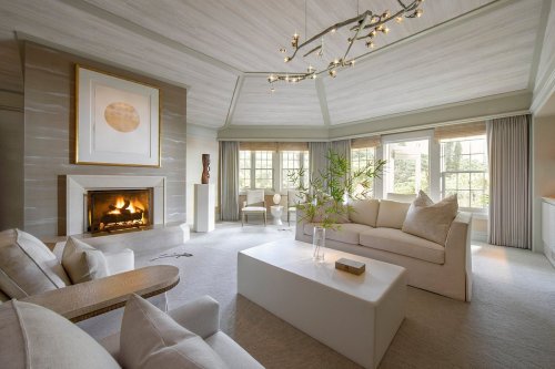 A modern living room with a fireplace, white furniture, large windows, and a stylish chandelier.