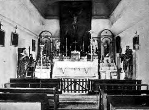 Interior of a church featuring an altar, statues, and wooden pews, with a crucifix prominently displayed.