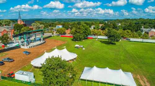 Aerial view of a grassy field with tents, a stage, and trees, set against a backdrop of blue skies and distant hills.