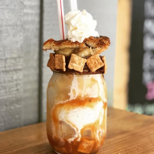 A decadent milkshake topped with whipped cream, pie crust, and cereal, served in a mason jar with two straws.