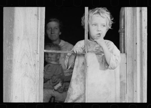 A young child stands by a window, looking outside while sucking their thumb; a woman and another child are in the background.