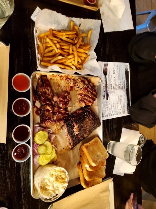A wooden table with a platter of ribs, fries, pickles, onions, toast, and four sauces in small cups.