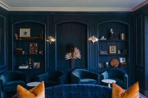 A stylish lounge with navy blue walls, plush chairs, decorative shelves, and a floral arrangement in a vase.