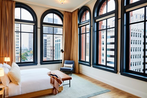 A stylish bedroom with large arched windows, city views, and modern furnishings, featuring a bed and a cozy chair.
