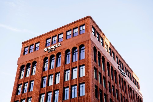 A corner view of the Shinola Hotel, featuring a brick facade and large windows against a clear blue sky.