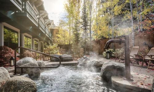 A serene outdoor hot spring surrounded by rocks, wooden chairs, and autumn foliage, with steam rising in the air.