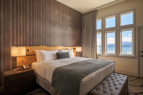 Cozy hotel room with a king bed, bedside lamps, and large windows showcasing a scenic view of the lake and mountains.