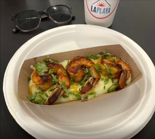 A paper tray with grilled shrimp on a bed of creamy sauce, garnished with green onions and cilantro, beside glasses and a drink.