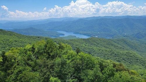 A panoramic view of lush green mountains and a winding river under a bright blue sky with fluffy white clouds.