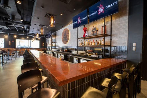 A modern bar with a polished countertop, shelves of liquor, and two screens displaying the Smoke Craft logo.