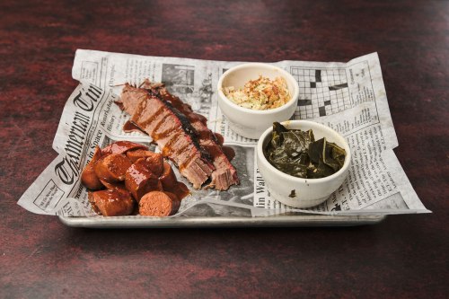 A plate of barbecue brisket, sausage, coleslaw, and collard greens served on newspaper.