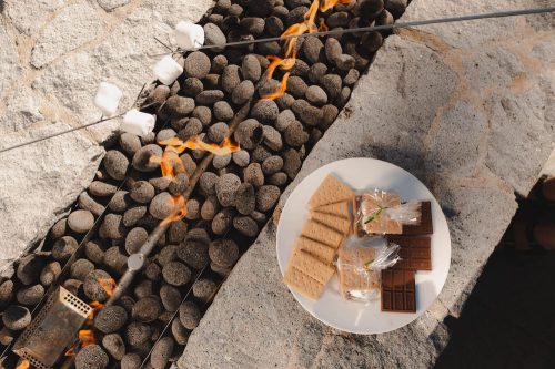 A plate with graham crackers, chocolate bars, and marshmallows near a fire pit with glowing embers.
