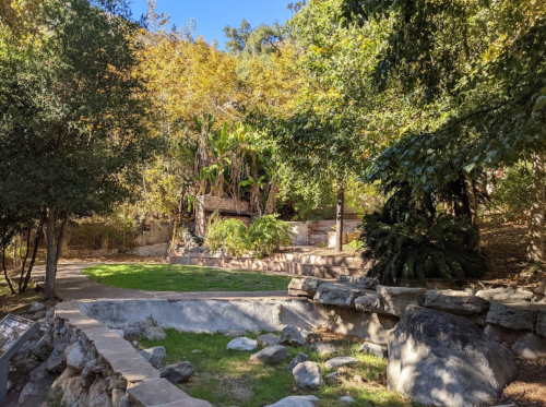 A serene park scene with lush greenery, stone pathways, and a small rocky area surrounded by trees.