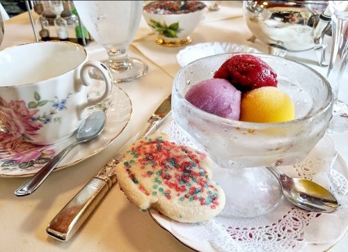 A glass dish with three colorful sorbet scoops beside a decorated cookie and a floral teacup on a table setting.